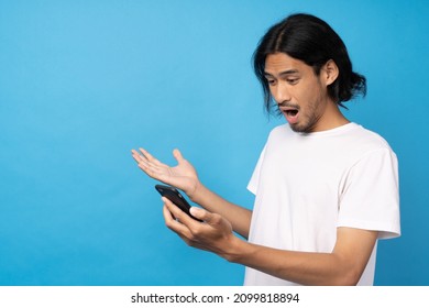 Cool Asian Man Excited And Surprised By Smartphone On Blue Background. A Man Saw A Sale On An Online Shopping Website Or See The Results Of The Football Team Cheering Competition.