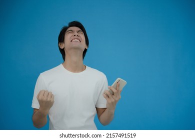 Cool Asian Man Excited And Surprised By Smartphone On Blue Background. A Man Saw A Sale On An Online Shopping Website Or See The Results Of The Football Team Cheering Competition.