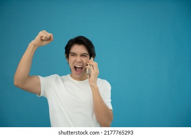Cool Asian Man Excited And Surprised By Smartphone On Blue Background. A Man Saw A Sale On An Online Shopping Website Or See The Results Of The Football Team Cheering Competition.