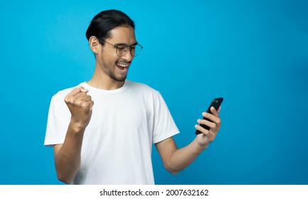 Cool Asian Woman Excited And Surprised By Smartphone On Blue Background. A Man Saw A Sale On An Online Shopping Website Or See The Results Of The Football Team Cheering Competition.