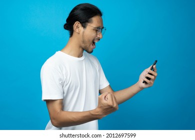 Cool Asian Woman Excited And Surprised By Smartphone On Blue Background. A Man Saw A Sale On An Online Shopping Website Or See The Results Of The Football Team Cheering Competition.