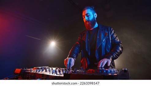 Cool bearded disc jockey working at mixer controller in a nightclub. Authentic dj performing in neon lights and smoke - nightlife concept 
