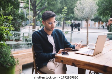 Cool Bearded Ethnic Guy In Casual Clothes And Eyeglasses Sitting With Legs Crossed And Taking Notes In Planner While Chilling With Laptop Cup Of Coffee And Smartphone On Street Cafe