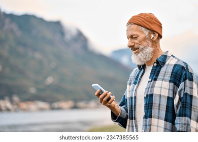 Cool bearded healthy older elder hipster man standing in nature park wearing earbud using mobile phone. Mature traveler enjoying freedom, active leisure lifestyle, listening music on smartphone. - Powered by Shutterstock