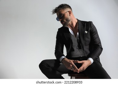 Cool Bearded Man In Tuxedo With Open Collar Shirt Looking To Side And Touching Fingers, Posing In A Stylish Way On Grey Background