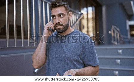Cool bearded young hispanic man caught in a serious phone conversation, standing on a sunny urban street, totally engrossed, seemingly undisturbed by city life bustle.