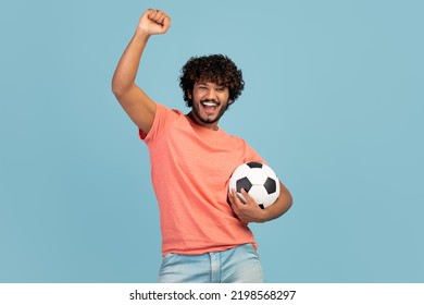 Cool Bearded Young Indian Man With Curly Hair Football Fan Holding Soccer Ball, Gesturing And Smiling, Watching Game, Blue Studio Background, Copy Space. Sports Betting, Gambling Concept