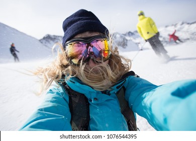 Cool, beautiful, young and attractive teenager or active lifestyle woman makes photo selfie on smartphone or action camera on ski slopes when snowboarding in mountains, wears goggles for snow and sun