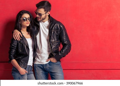 Cool beautiful young couple in leather jackets and sunglasses standing against red background. Man is looking at his woman