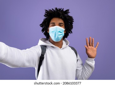 Cool Black Teen Guy In Medical Face Mask Taking Selfie And Waving At Camera On Violet Studio Background. Funky African American Teenager Making Photo Of Himself, Wearing Facial Anti Covid Protection