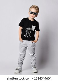 Cool Blond Kid Boy In Sunglasses, Black T-shirt With Dinosaur Print And Gray Pants Stands Leaning Sideways Holding Hands In Pockets Over Gray Background