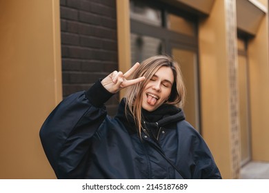 Cool Blonde Woman In Black Hoodie And Navy Blue Bomber Shows Peace Sign, Show Tongue, Close Eyes And Posing Outside. Happy Woman Walk And Posing Outdoor.