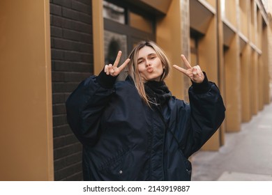 Cool Blonde Woman In Dark Blue Bomber Shows Peace Sign, Blows Kiss And Posing Outside. Happy Woman Walk And Posing Outdoor.