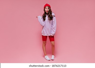 Cool Brunette Girl In Red Hat, Cycling Shorts And Purple Hoodie Whistles. Curly Woman With Bright Lips Poses On Pink Isolated Background.