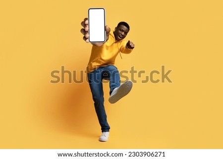 Cool cheerful handsome young black guy hipster showing smartphone with white blank screen, thumb up and shoe sole, isolated on yellow studio background, mockup. Digital life, modern technologies