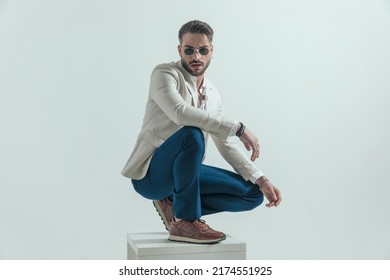 Cool Crouched Man With Glasses Holding Elbow On Knee In A Fashion Way And Posing On Grey Background In Studio