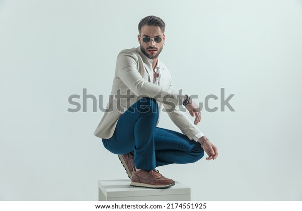 cool crouched man\
with glasses holding elbow on knee in a fashion way and posing on\
grey background in studio