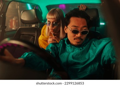A cool diverse couple is bonding in the car. The man is sitting in the drivers seat and the woman is in the backseat with a lollypop looking over his shoulder.