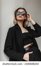 Cool Elegant Fashion Young Woman With Stylish Eyewear In Black Fashionable Business Outfit With Top And Blazer Poses Near A White Wall On The Street