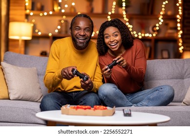 Cool emotional happy african american couple in casual outfit sitting on couch in festive living room, holding joysticks and laughing, playing video games at home, eating pizza - Powered by Shutterstock