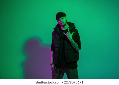 Cool fashionable hipster man with a hat in stylish black clothes with a vest posing in a creative color studio with green and purple light - Shutterstock ID 2278075671