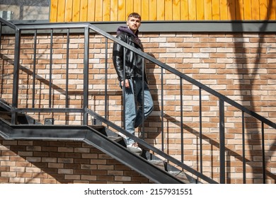 Cool Fashionable Man In Stylish Spring Clothes With A Jacket, Hoodie And Jeans Walks Down The Stairs Outside A Brick Building