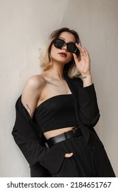 Cool fashionable vogue business woman in black stylish clothes with top, blazer and pants wears a fashion sunglasses and stands near a white wall