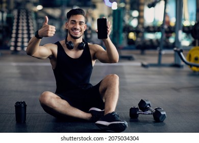 Cool Fitness App. Young Arab Man Posing In Gym With Blank Smartphone In Hands, Smiling Middle Eastern Male Athlete Sitting On Floor, Holding Cellphone With Blank Screen And Showing Thumb Up, Mockup - Powered by Shutterstock