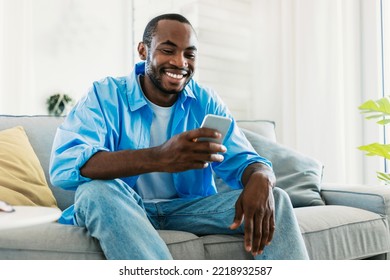 Cool gadget and app concept. Portrait of happy black man using mobile phone, watching video while sitting on sofa in living room, free space. Guy browsing internet on cellphone - Powered by Shutterstock