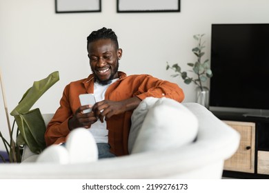 Cool Gadget And App. Excited Black Man Holding Mobile Phone And Typing Sms Message, Resting On Sofa At Home, Free Space. Man Browsing Internet And Surfing Web