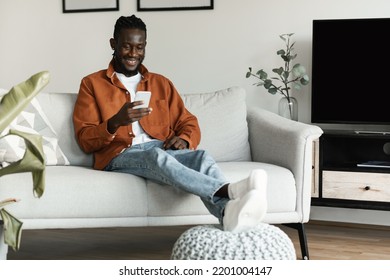 Cool Gadget And Application. Happy Black Man Holding Cellphone, Typing Sms Message, Sitting On The Sofa In Living Room, Copy Space. Guy Surfing Web, Using App