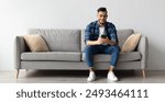 Cool Gadget And Application. Portrait of young smiling Arab man holding mobile phone, typing sms message, sitting on the couch in living room. Guy browsing internet, surfing web, using app, free space