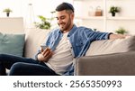 Cool Gadget And Application. Portrait of young smiling jewish man holding mobile phone, typing sms message, sitting on comfy sofa in living room. Guy resting browsing internet, surfing web, using app