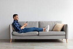 Cool Gadget And Application. Portrait Of Young Smiling Arab Man Holding Mobile Phone, Typing Sms Message, Sitting On The Sofa In Living Room. Guy Browsing Internet, Surfing Web, Using App, Free Space
