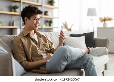 Cool Gadget And Application. Portrait Of Young Smiling Asian Man Holding Mobile Phone, Typing Sms Message, Sitting On The Sofa In Living Room. Guy Browsing Internet, Surfing Web, Using App, Free Space