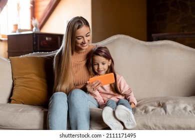 Cool Gadget And Application. Portrait Of Young Smiling Woman And Little Girl Using Mobile Phone, Sitting On Couch In Living Room. Family Browsing Internet, Watching Cartoon Together, Free Copy Space