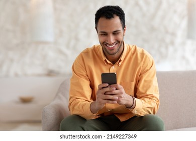 Cool Gadget And Application. Portrait Of Young Smiling Arab Man Holding Mobile Phone, Typing Sms Message, Sitting On The Couch In Living Room. Guy Browsing Internet, Surfing Web, Using App, Free Space