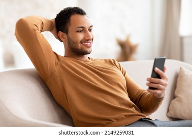 Cool Gadget And Application. Portrait Of Young Smiling Arab Man Using Mobile Phone, Watching Video, Sitting On The Couch In Living Room Leaning Back Holding Hand Behind Head. Guy Browsing Internet