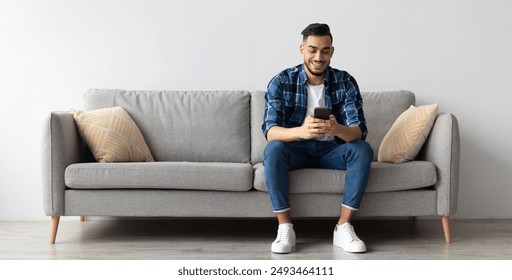 Cool Gadget And Application. Portrait of young smiling Arab man holding mobile phone, typing sms message, sitting on the couch in living room. Guy browsing internet, surfing web, using app, free space - Powered by Shutterstock