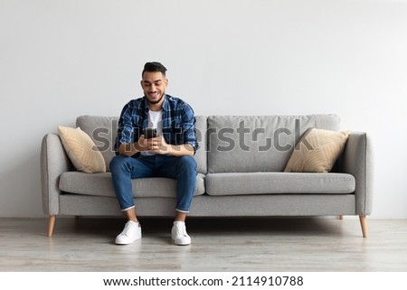 Cool Gadget And Application. Portrait of young smiling Arab man holding mobile phone, typing sms message, sitting on the couch in living room. Guy browsing internet, surfing web, using app, free space