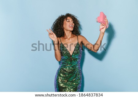 Cool girl with fluffy wavy hairstyle in green stylish dress showing tongue and peace sign, holding pink camera and making selfie on blue background..