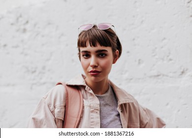 Cool girl with short hair looking into camera on background on white backdrop. Brunette lady with glass in beige outside posing on backdrop of wall..