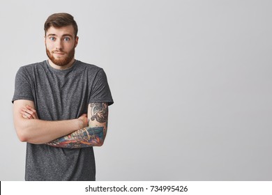 Cool good-looking young bearded man with stylish hairstyle and tattooed arm crossing hands with unsatisfied expression showing friend he have no idea what to do with situation.