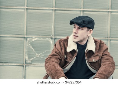 Cool Guy In Aviator Jacket And Newsie Cap Relaxes Against A Glass Wall