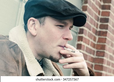 Cool Guy In Aviator Jacket And Newsie Cap Relaxes Against A Glass Wall And Enjoys His Cigarette