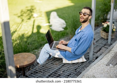 Cool Guy Works On Laptop While Sitting At Green Lawn Of Country House Backyard. Concept Of Remote Work At Cozy Home Workplace And Freelance