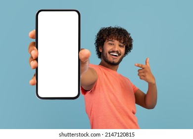 Cool handsome curly young indian guy in stylish pink t-shirt showing brand new cell phone with white empty screen and gesturing, sharing exciting online deal, blue background, mockup - Shutterstock ID 2147120815