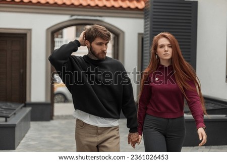 Cool handsome fashion brutal hipster man with a beard and hairstyle in a black pullover and a beautiful red-haired stylish girl model with a red hoodie walks on the street