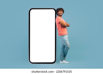 Cool handsome millennial hindu guy leaning on big cell phone with white empty screen, holding arms crossed, blue studio background, mockup for mobile app or advertisement, copy space - Shutterstock ID 2206791535
