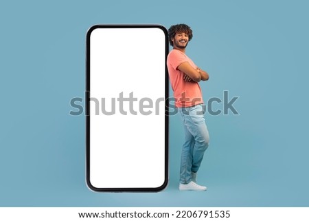 Cool handsome millennial hindu guy leaning on big cell phone with white empty screen, holding arms crossed, blue studio background, mockup for mobile app or advertisement, copy space